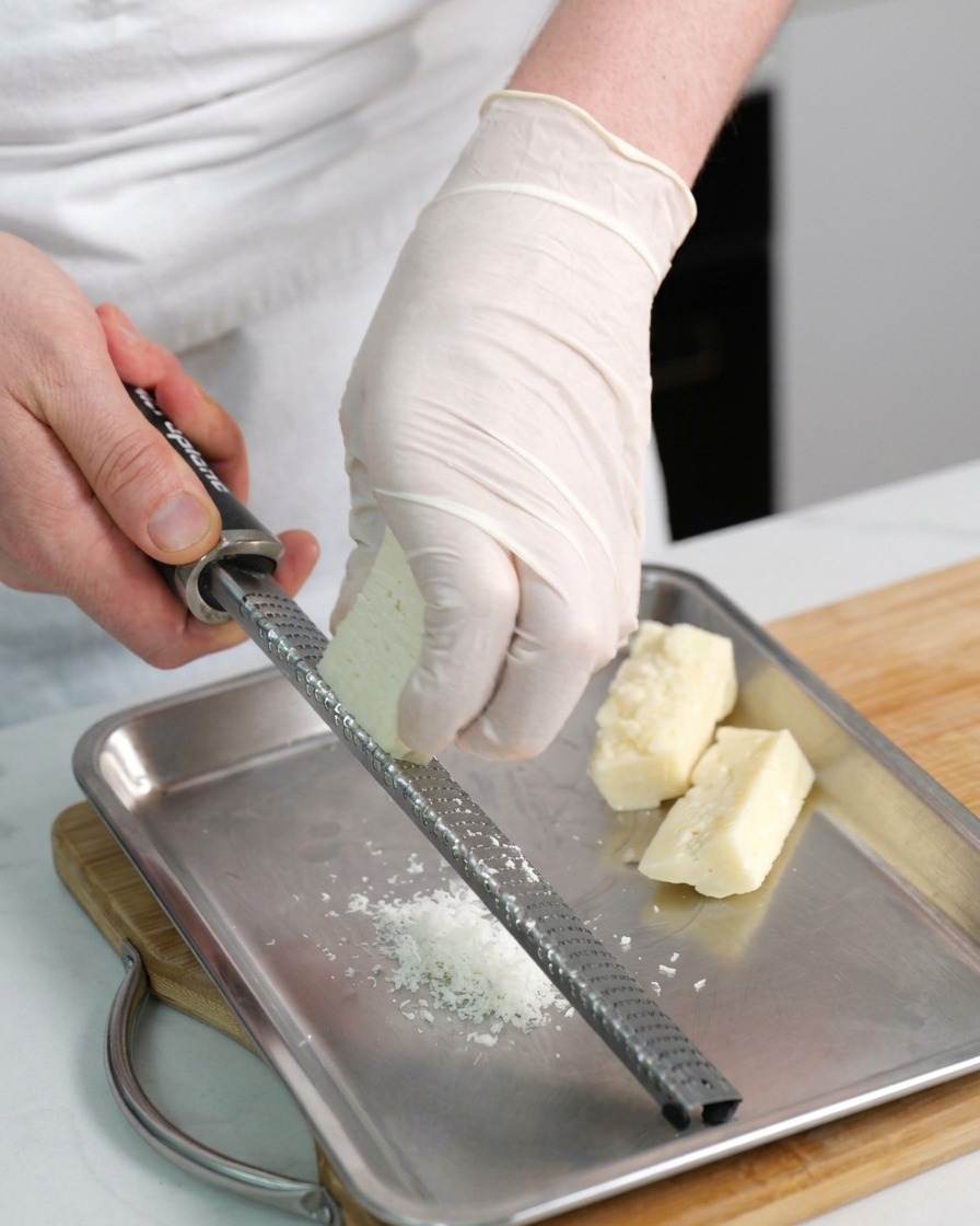 Mani che utilizzano una grattugia a mano per ridurre in scaglie un pezzo di Pecorino Romano su un vassoio di metallo.
