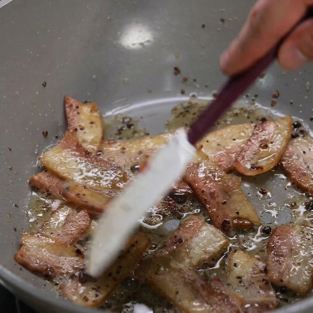 Strisce di guanciale che rosolano in padella mentre il grasso si scioglie, mescolate con una spatola chiara.