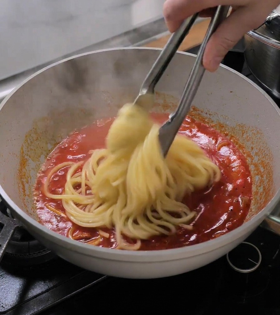 Pinze da cucina che trasferiscono i bucatini cotti direttamente nella padella con il sugo di pomodoro.