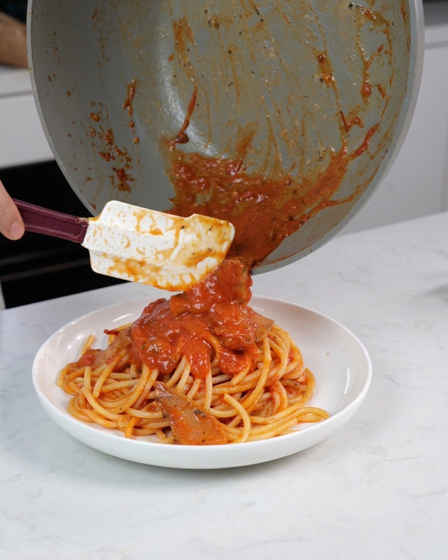 Una mano che utilizza una spatola per raccogliere il sugo e i pezzi di carne dalla padella e versarli sui bucatini già impiattati.