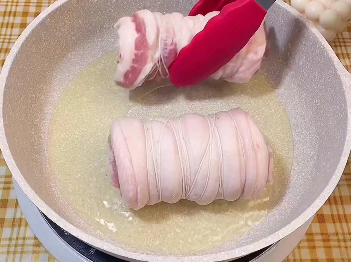 Pinze rosse che adagiano il rotolo di maiale legato in una padella sfrigolante con olio.