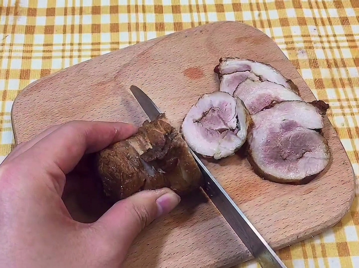 Mano che usa un coltello per affettare un rotolo di chashu in fette sottili su un tagliere di legno.