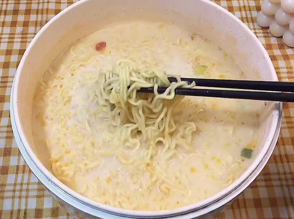 Bacchette nere che sollevano una porzione di noodles ondulati da un brodo cremoso.