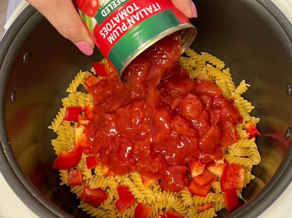 Una mano che versa una lattina di pomodori a cubetti sopra la pasta e i peperoni.