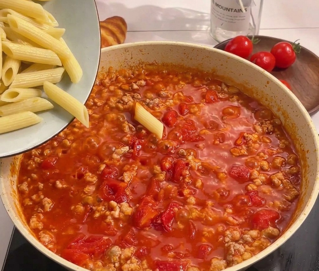Penne cotte versate da una ciotola in una padella con sugo di pomodoro e carne.