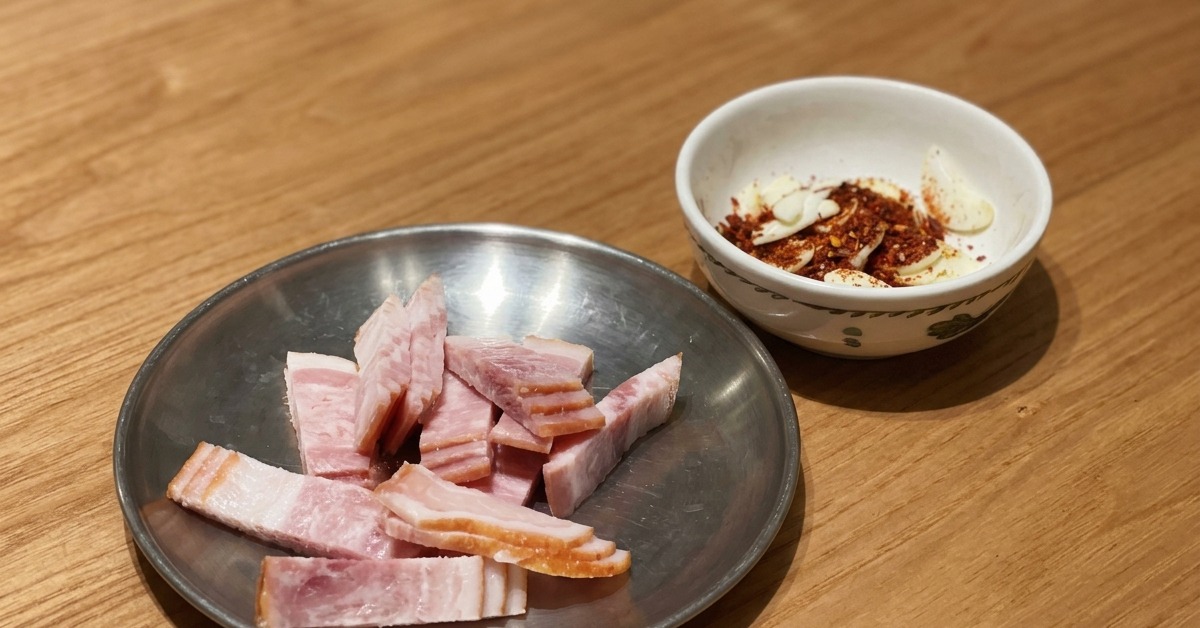 Pancetta cruda a fette su un piatto di metallo accanto a una ciotolina con aglio affettato e peperoncino su un tavolo di legno.