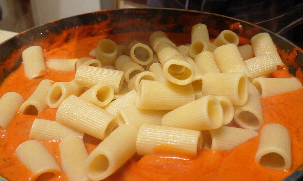 Rigatoni cotti aggiunti in una padella con salsa arancione-rossastra vellutata.