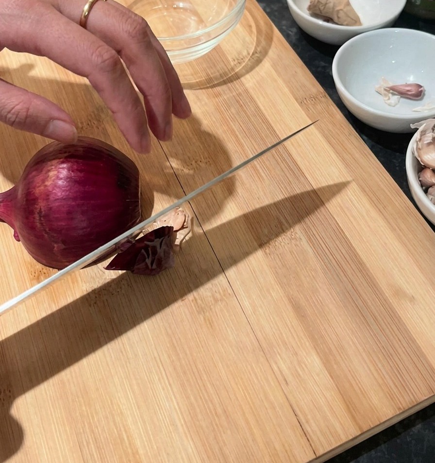 Una mano che tiene una cipolla rossa su un tagliere di legno con un coltello da chef pronto per affettare.