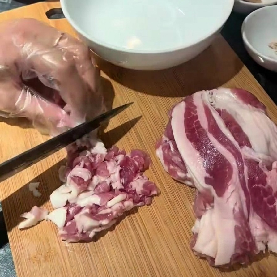 Una mano guantata che usa un coltello nero per tagliare strisce di bacon crudo su un tagliere di legno.