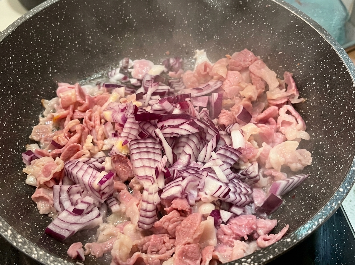 Cipolla rossa tritata sparsa sopra i pezzi di bacon parzialmente cotti in una padella calda.