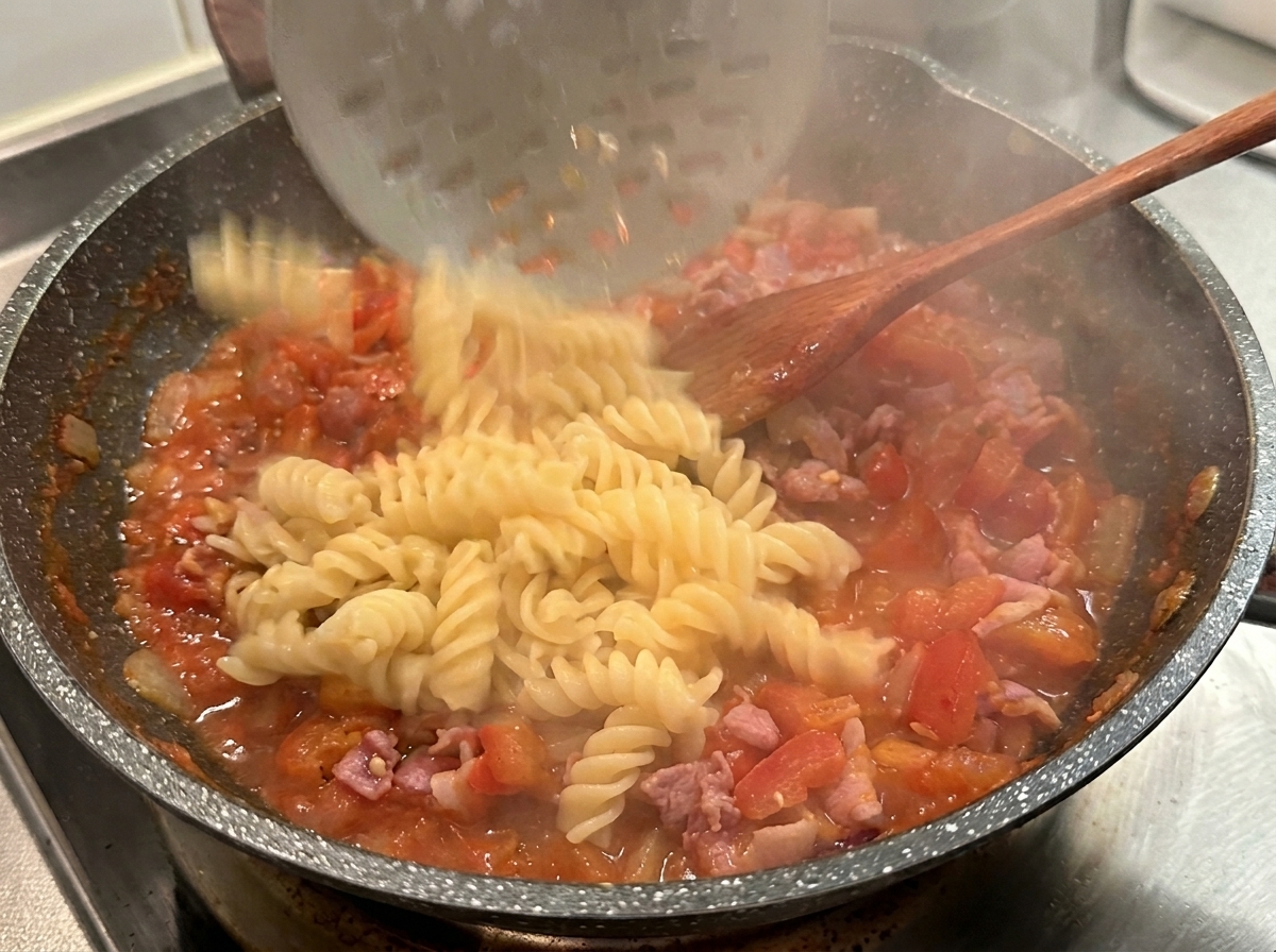 Fusilli cotti versati da un mestolo forato in una padella calda con sugo di pomodoro e bacon.