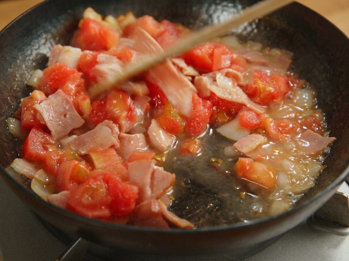 Spatola che mescola pomodori, bacon e cipolle in una padella nera.
