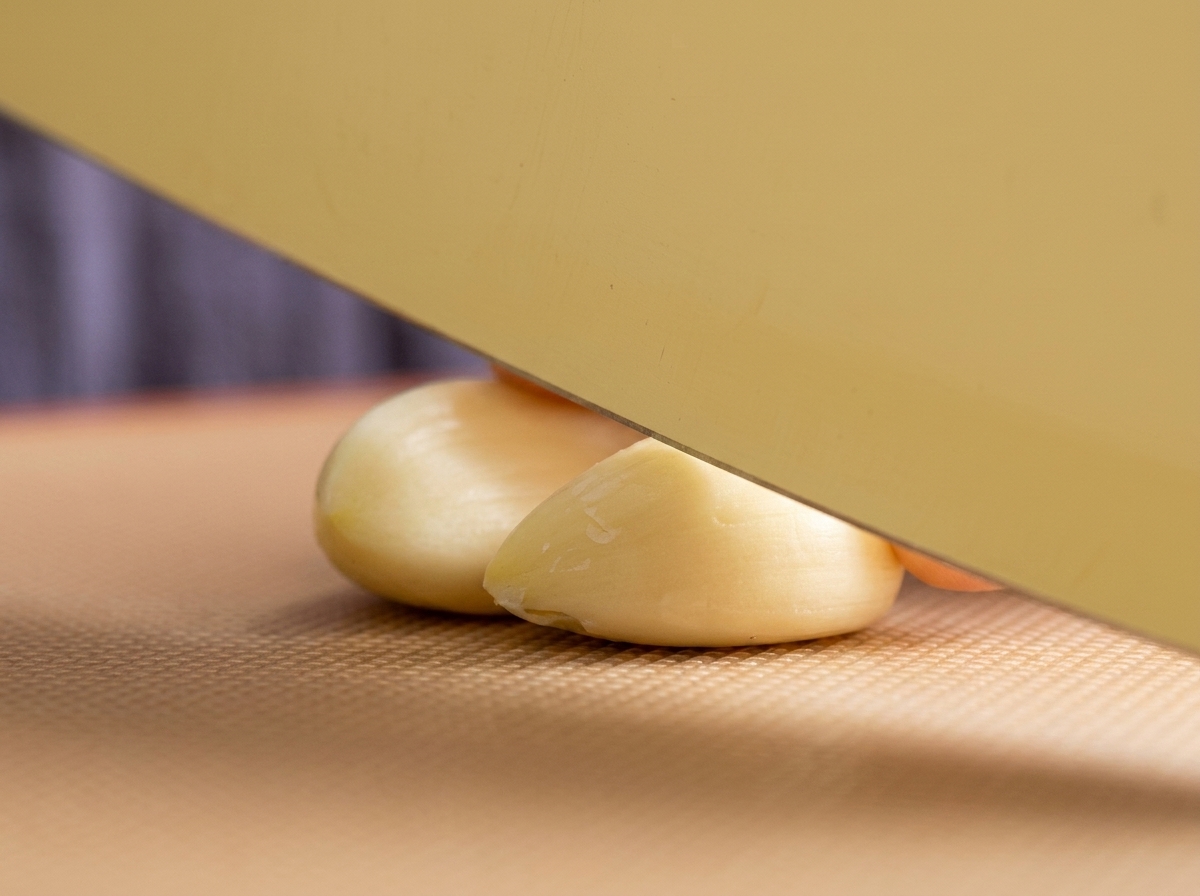 Un coltello da cucina appoggiato su due spicchi daglio sbucciati su un tagliere marrone.