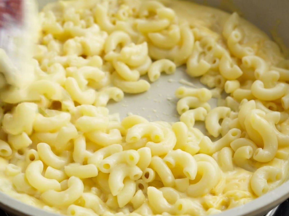 Una spatola che salta i maccheroni in padella, coprendoli completamente con la salsa al formaggio.