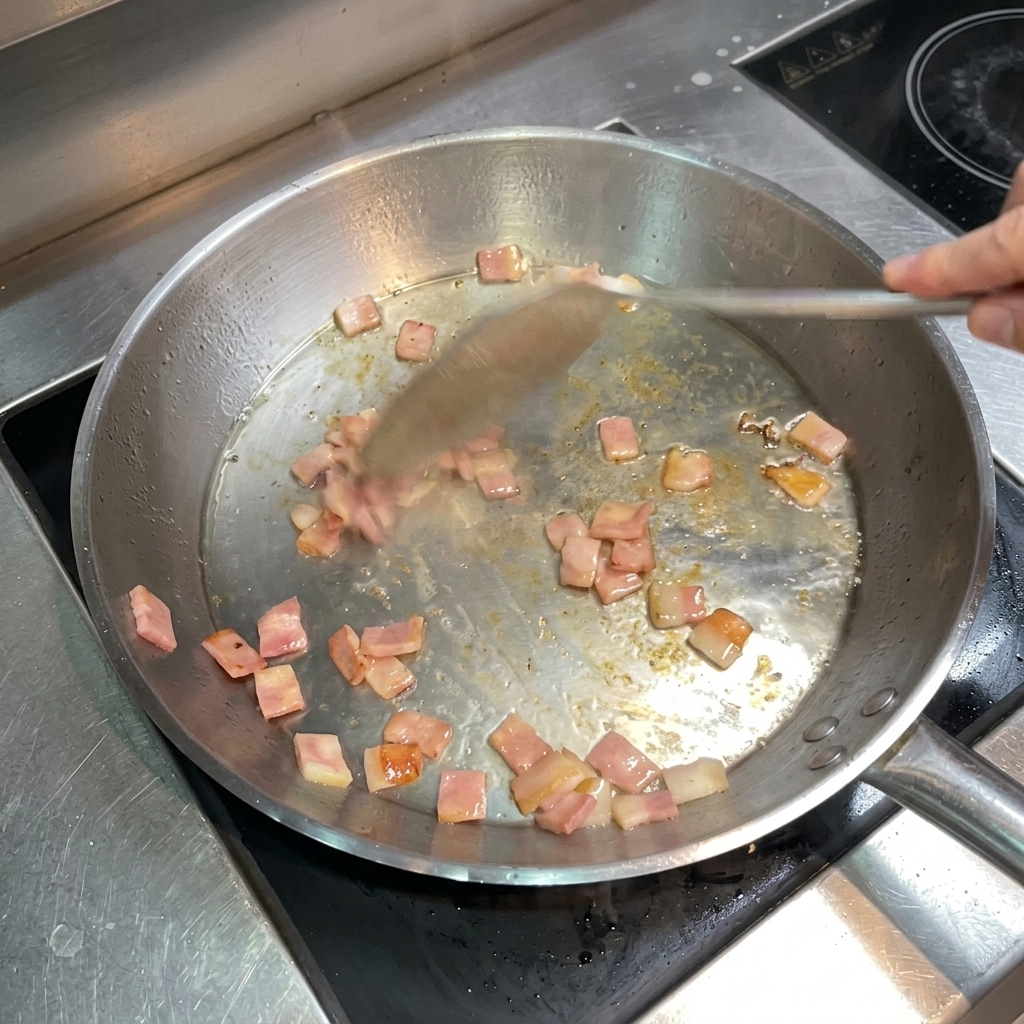 Una spatola di metallo che mescola pancetta a cubetti dorata e croccante in una padella.