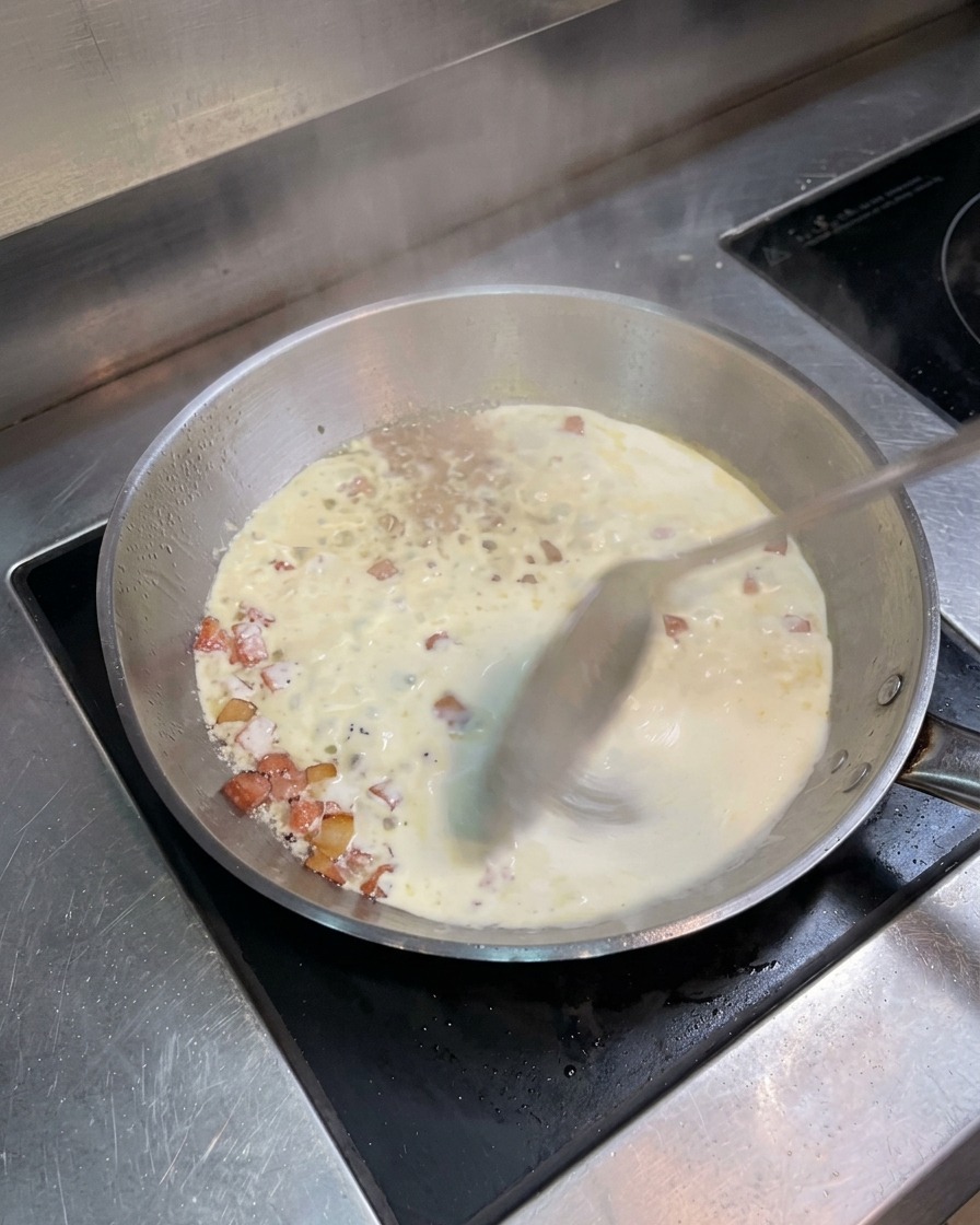 Una padella in acciaio inossidabile con panna e pancetta che sobbolle, mescolata con un cucchiaio di metallo.
