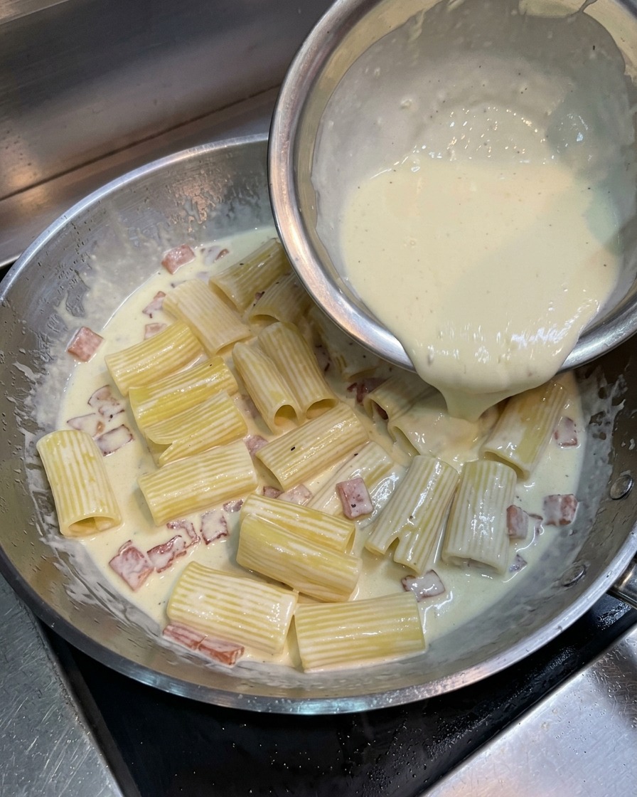 Versamento di un composto giallo di tuorlo, panna e formaggio sui rigatoni e pancetta in padella.