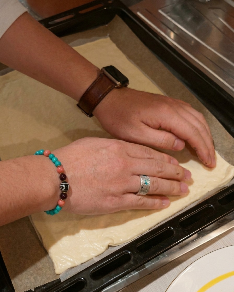 Mani che premono delicatamente i bordi di un impasto rettangolare per pizza su una teglia da forno scura.