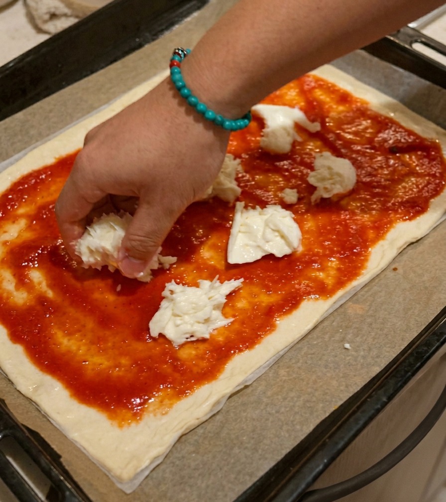 Una mano che distribuisce pezzi di mozzarella sfilacciata a mano su una pizza già condita con pomodoro.