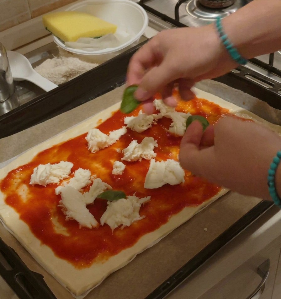 Mani che dispongono foglie intere di basilico fresco su una pizza cruda con pomodoro e mozzarella.