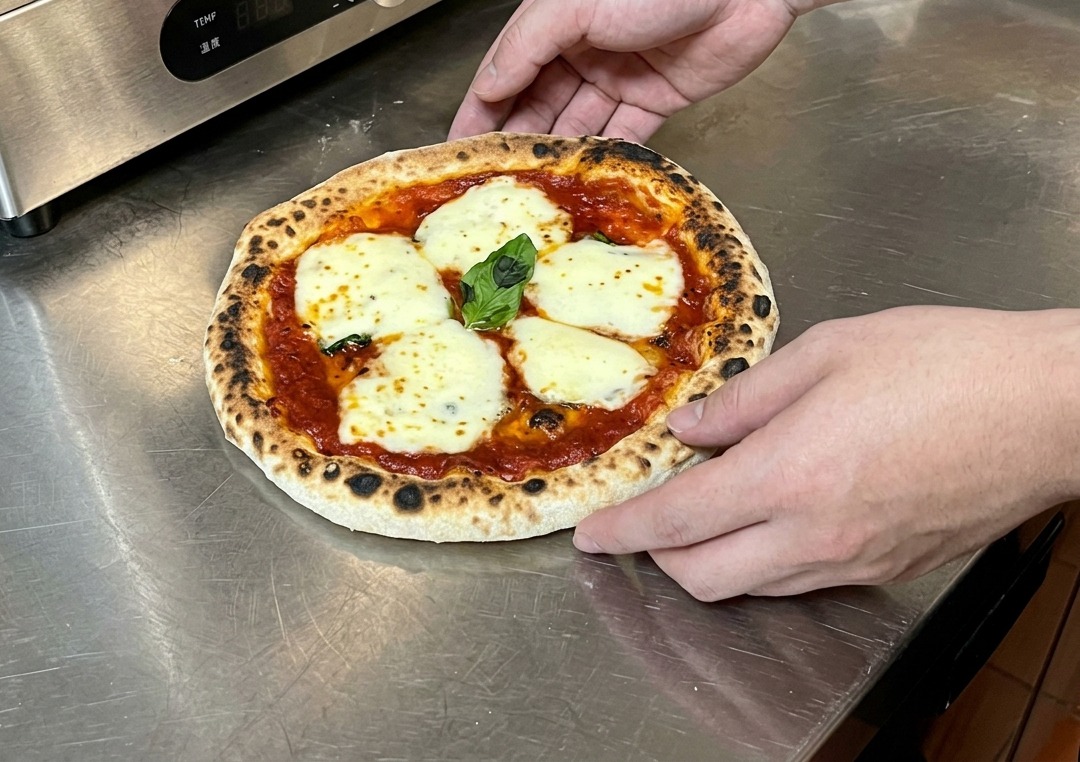 Mani che tengono una pizza appena sfornata su un piano di metallo, controllando la croccantezza della base.