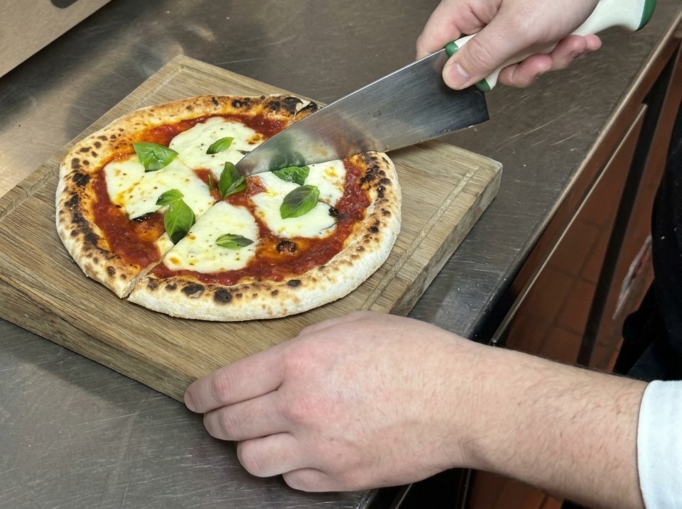 Mani che usano un grande coltello per tagliare una pizza Margherita a spicchi su un tagliere.