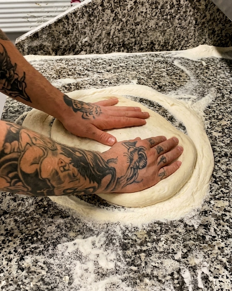 Mani tatuate che premono e stendono delicatamente un panetto tondo di pizza su una superficie di marmo infarinata.