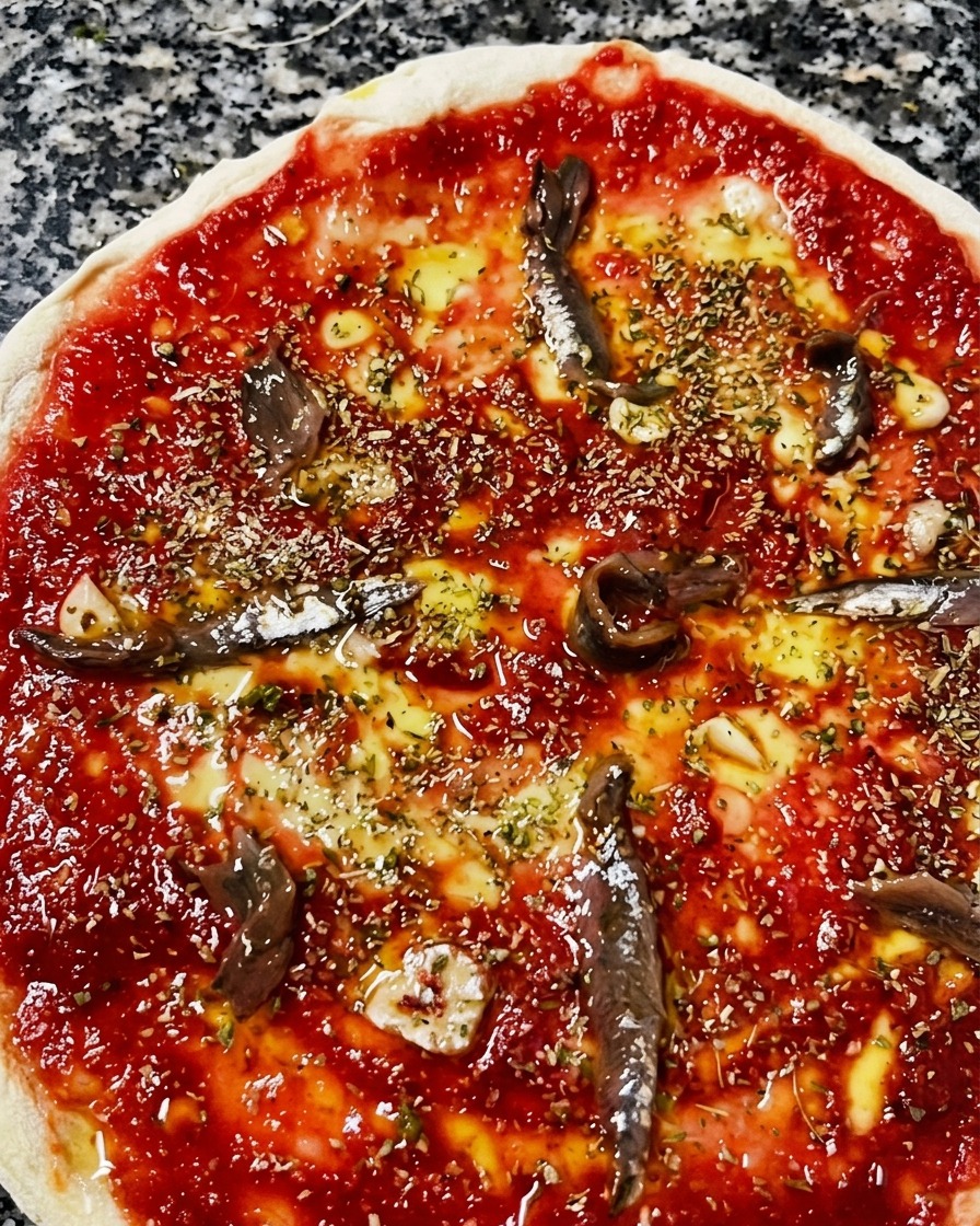 Vista ravvicinata dallalto di una pizza cruda con pomodoro, olio, origano, aglio e filetti di acciughe marinate.
