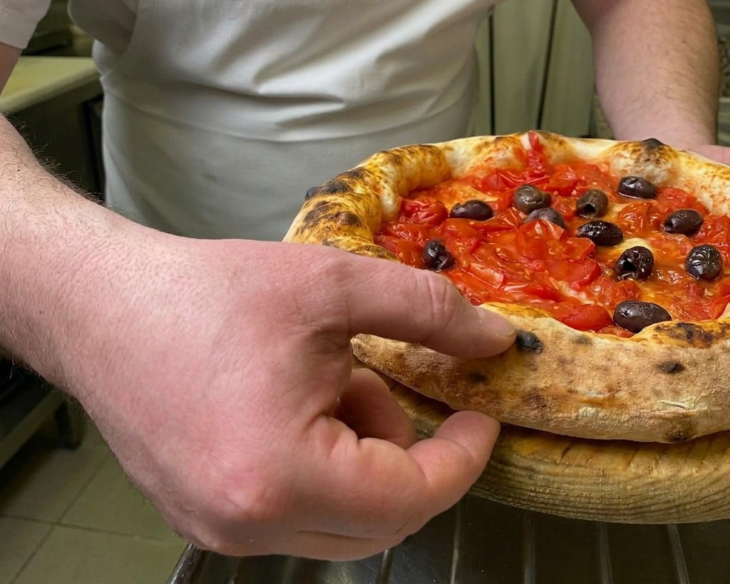 Uno chef che preme un dito sul cornicione alto e maculato di una pizza calda appena sfornata per testarne la sofficità.