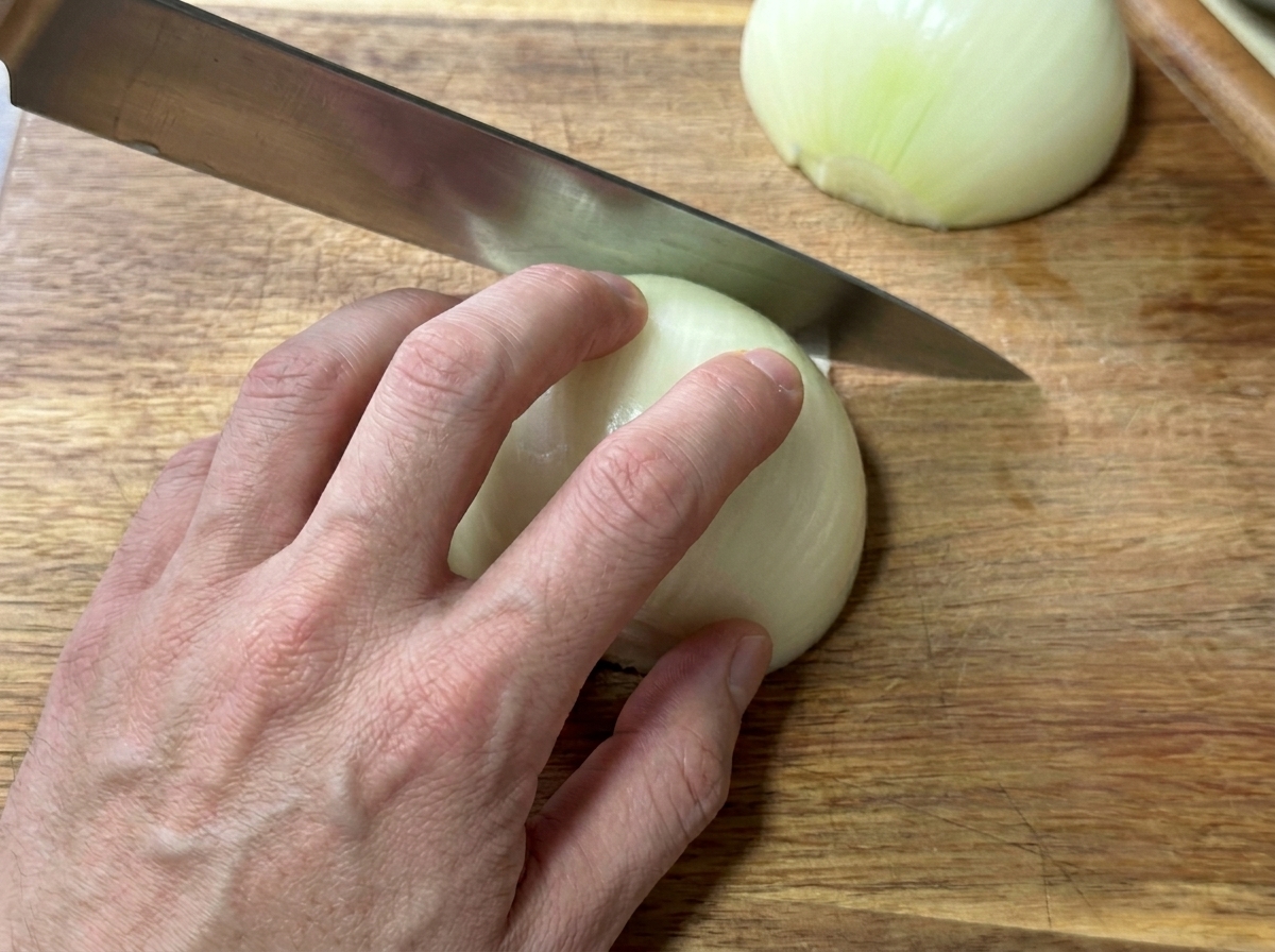 Una mano che affetta una cipolla bianca sbucciata in strisce sottili su un tagliere di legno.