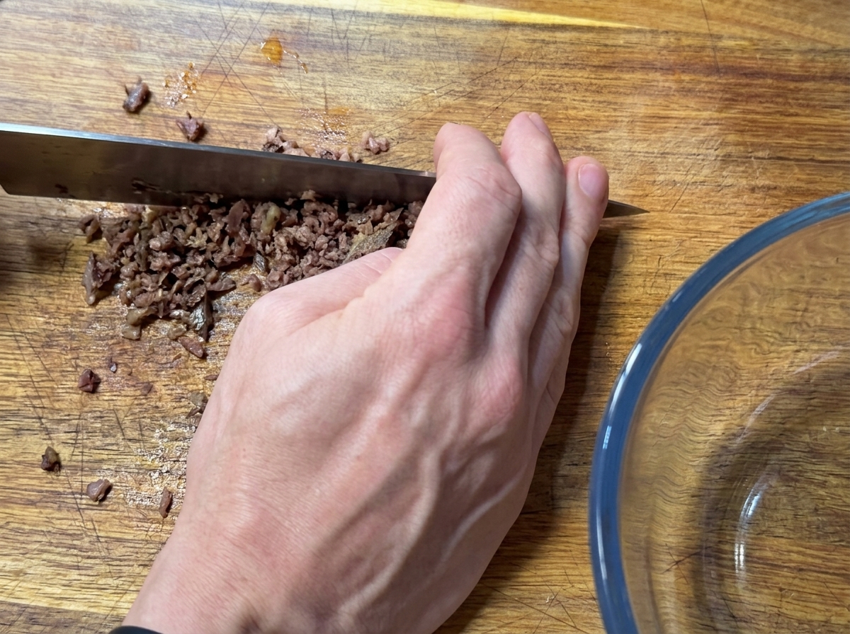 Una mano che usa un grosso coltello per tritare finemente filetti di acciughe su un tagliere.