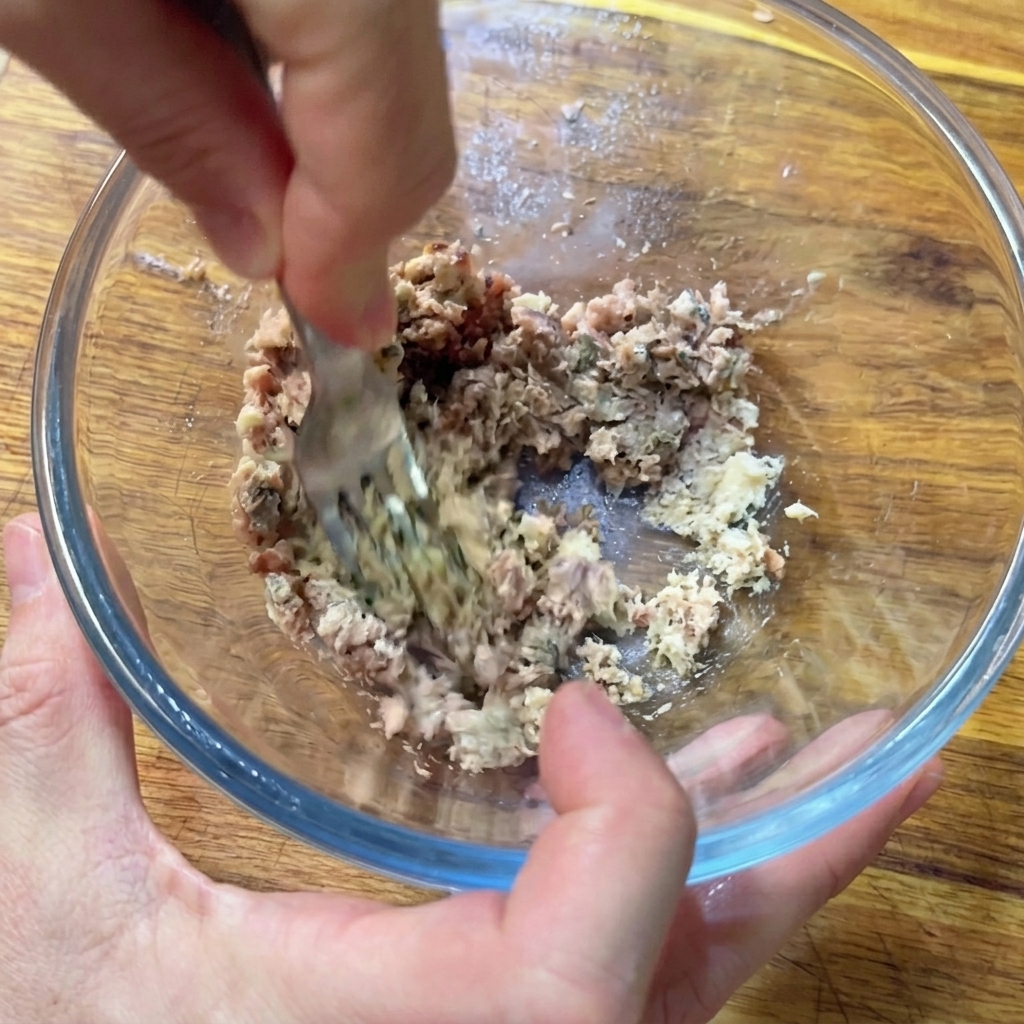 Mani che usano una forchetta per schiacciare acciughe e sardine in una pasta in una ciotola di vetro.