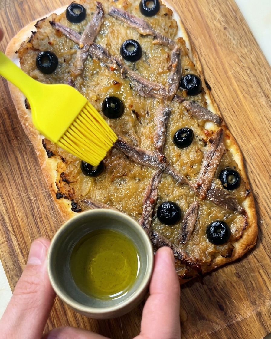 Una mano che spennella olio doliva su una Pissaladière appena sfornata.