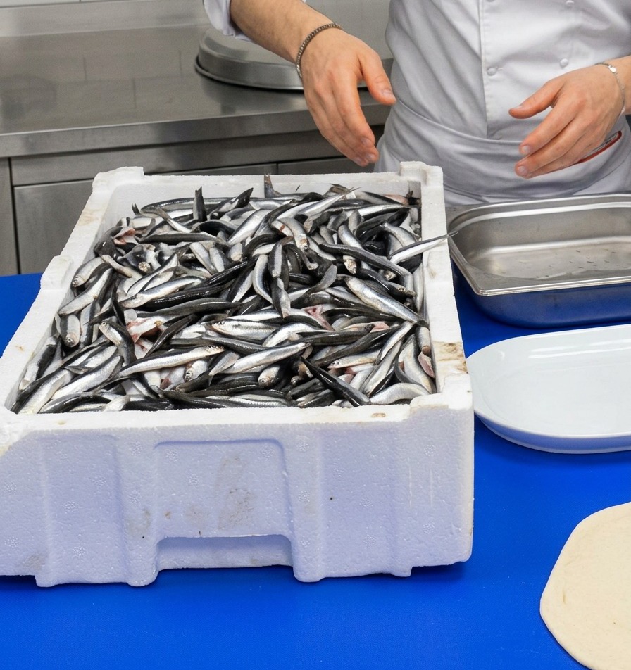 Uno chef in divisa bianca mentre maneggia una cassetta di polistirolo piena di alici fresche e argentee.