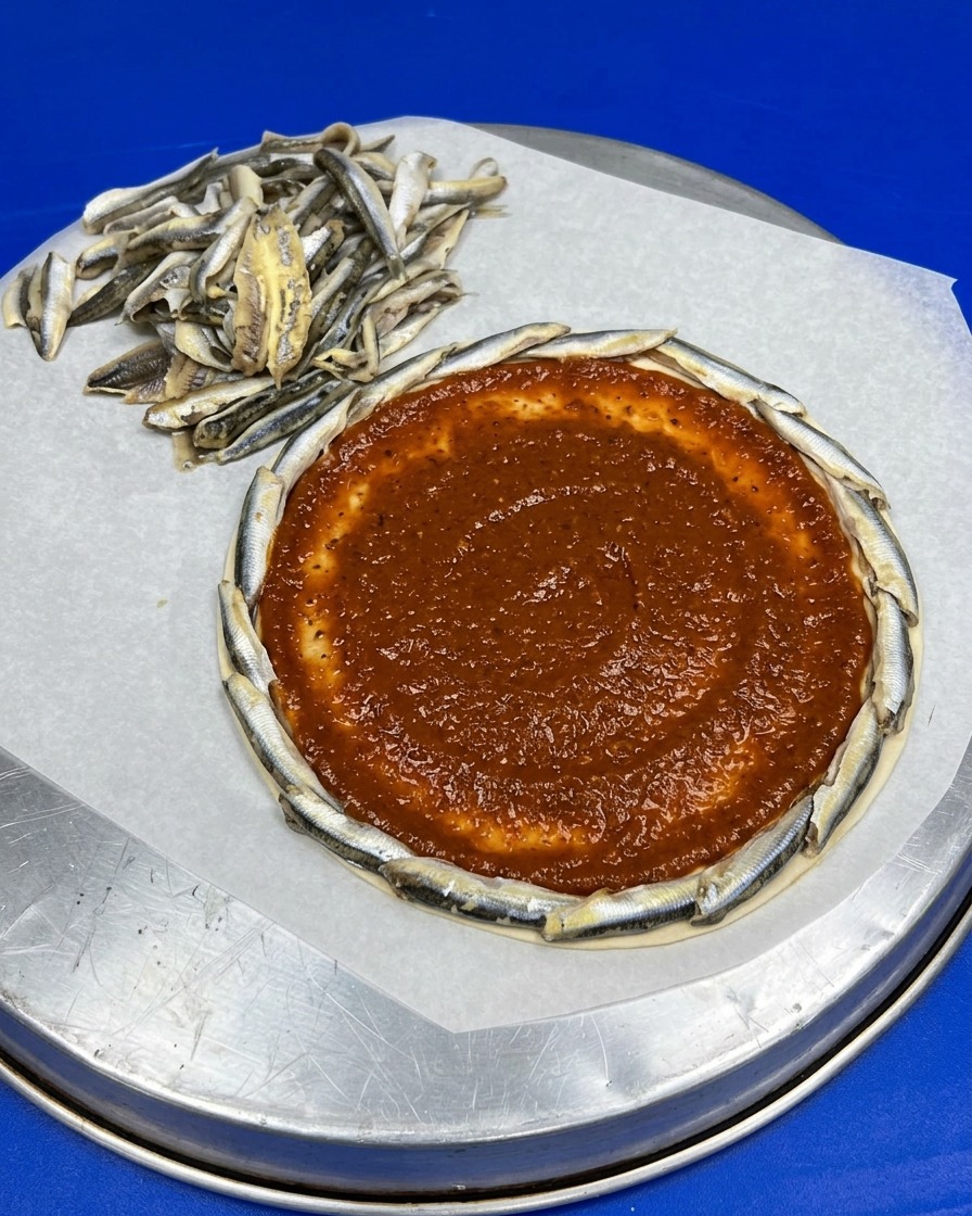 Una base pizza rotonda con bordo di alici e uno strato uniforme di salsa di pomodoro rossa stesa al centro.