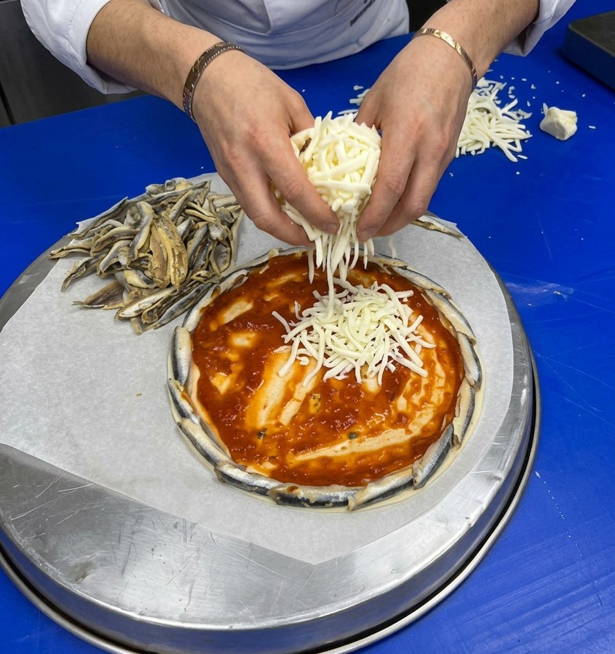 Mani che cospargono formaggio bianco grattugiato sopra la base di pomodoro circondata dal bordo di alici.