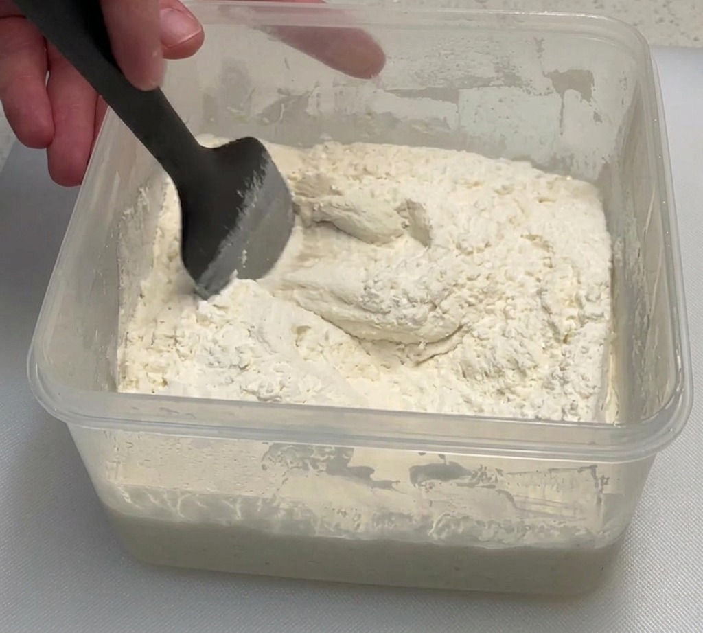 Una mano usa una spatola nera per mescolare farina e poolish in un contenitore trasparente.