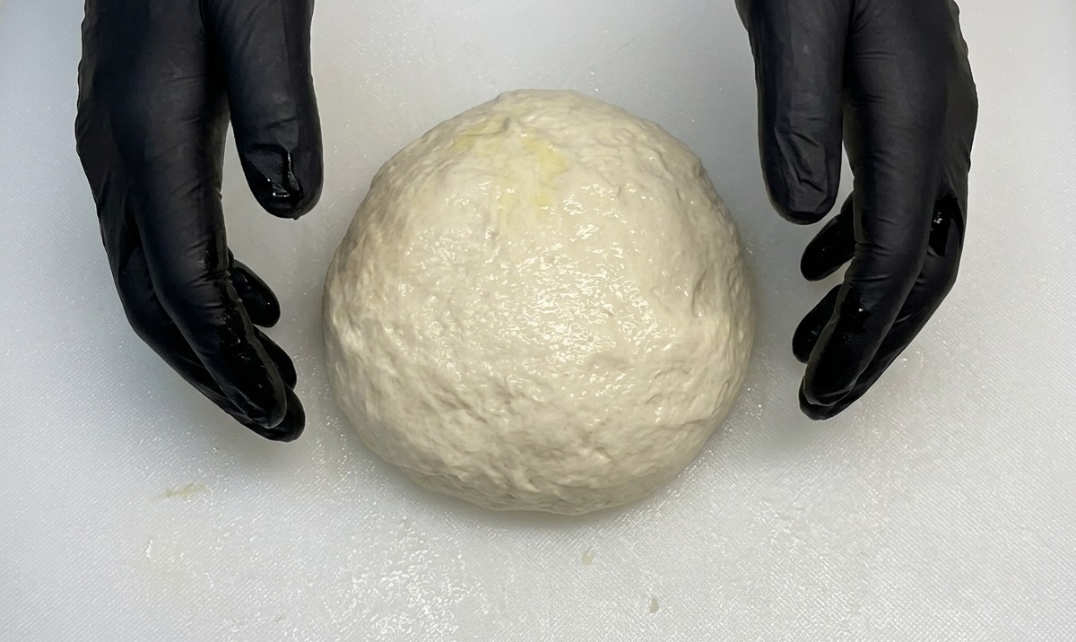 Mani guantate che modellano una palla di impasto per pizza liscia e rotonda su un piano bianco.