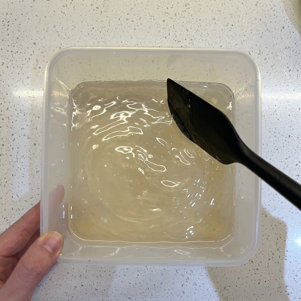 Una spatola nera che mescola farina, acqua e lievito in un contenitore di plastica trasparente.