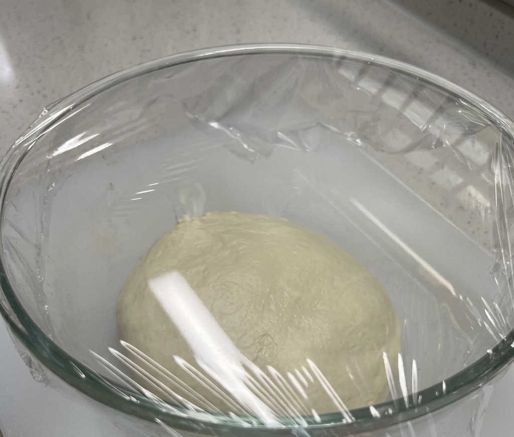 Una palla di impasto per pizza in una ciotola di vetro coperta da pellicola trasparente.