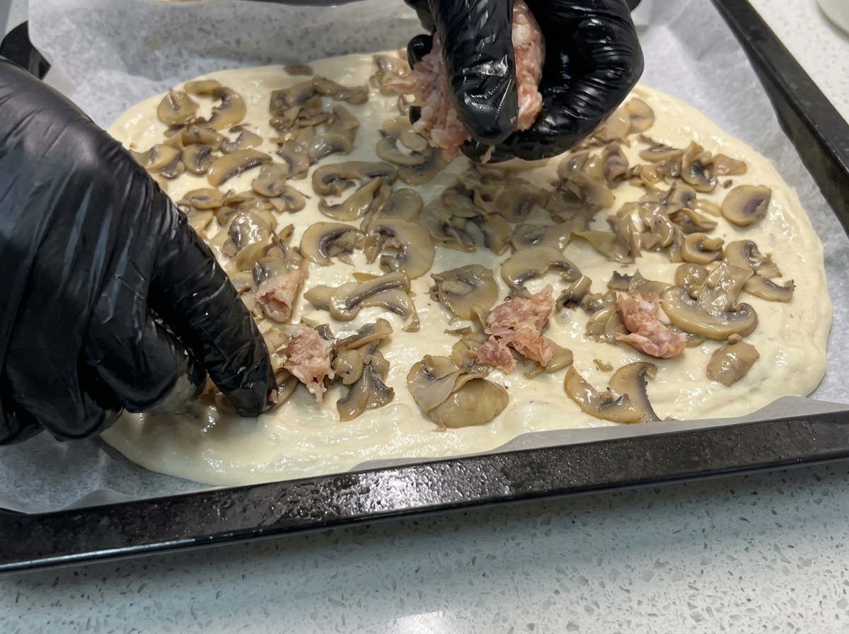 Mani che distribuiscono pezzi di salsiccia cruda sopra i funghi sulla base della pizza.