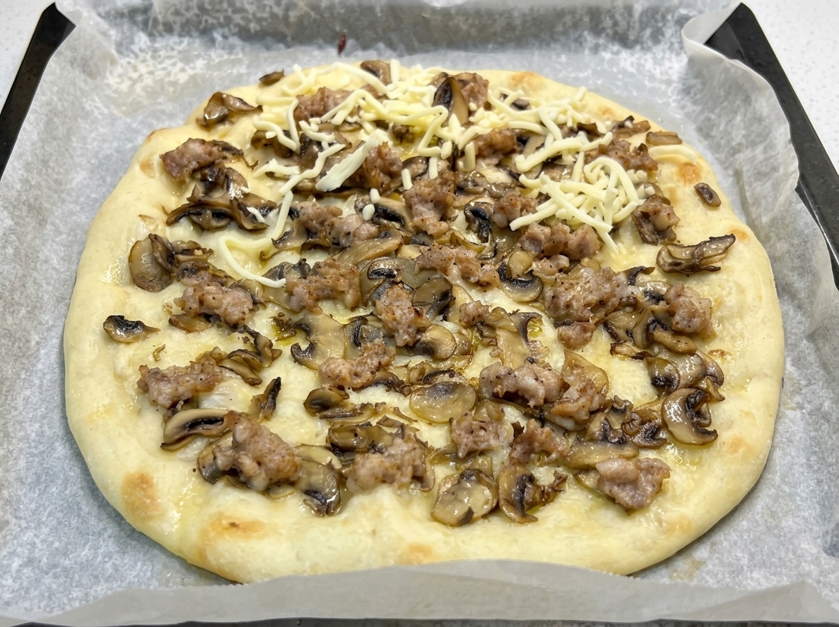 Pizza parzialmente cotta con salsiccia e funghi, ora cosparsa di mozzarella grattugiata.