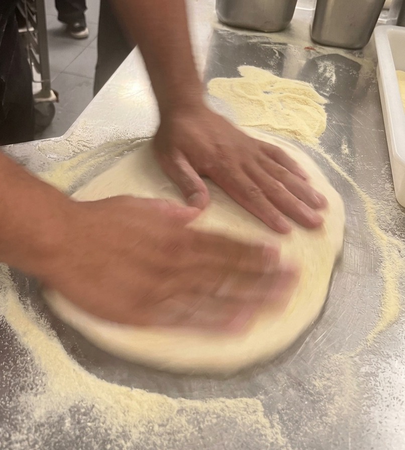 Mani di un cuoco che allargano limpasto per pizza fino a formare un cerchio su un bancone dacciaio.