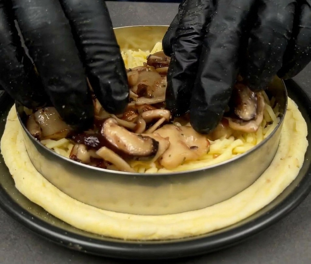 Mani con guanti neri che posizionano funghi misti cotti sopra un letto di mozzarella su una base per pizza.