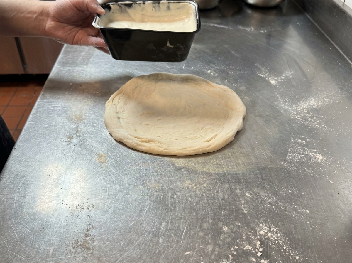 Salsa al tartufo versata su un disco di impasto per pizza su un tavolo dacciaio.