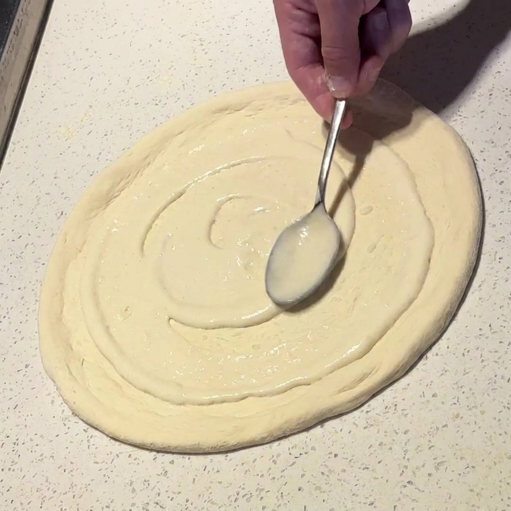 Un cucchiaio di metallo che stende una salsa bianca cremosa con movimento circolare sulla base della pizza.