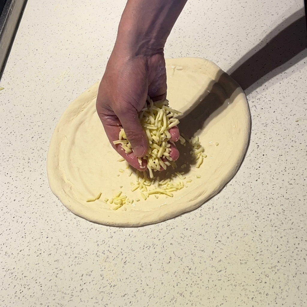 Una mano che cosparge mozzarella grattugiata sulla base della pizza con salsa bianca.