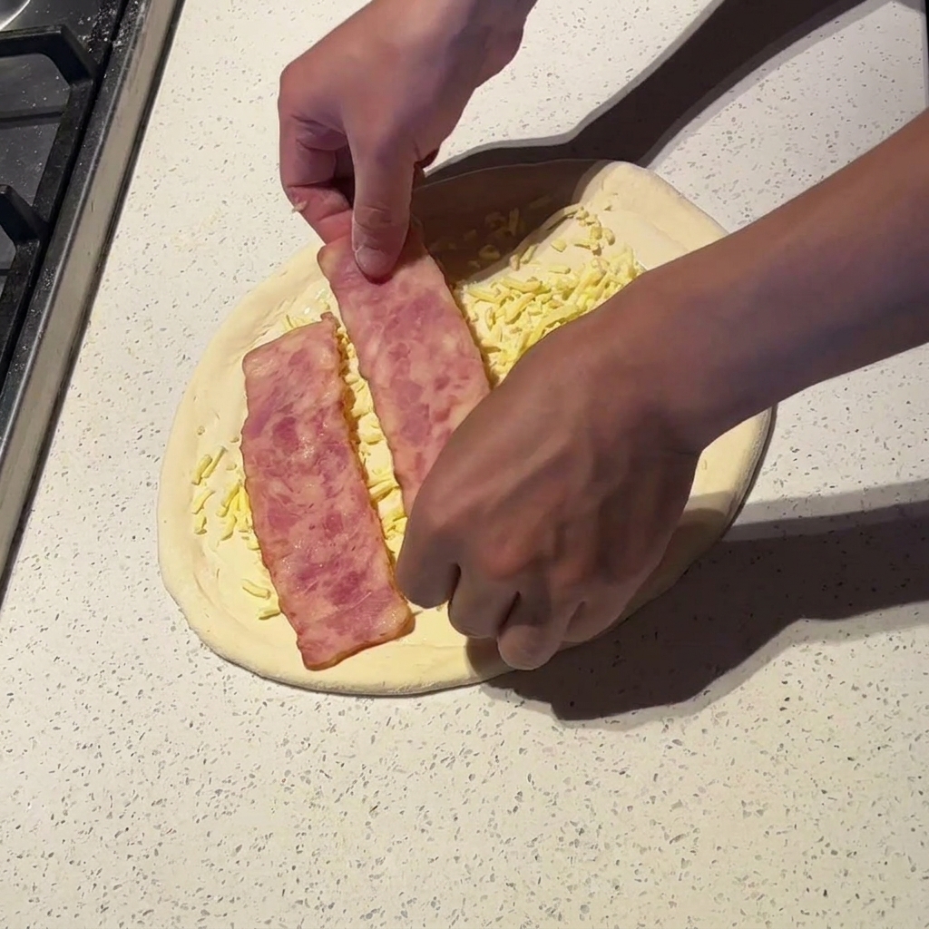 Mani che posizionano fette larghe di pancetta precotta su una pizza con salsa e formaggio.