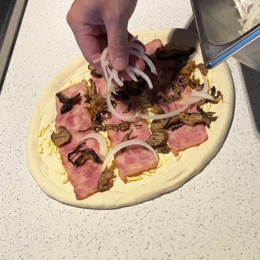 Una mano che sparge fette sottili di cipolla bianca su una pizza con pancetta e funghi.