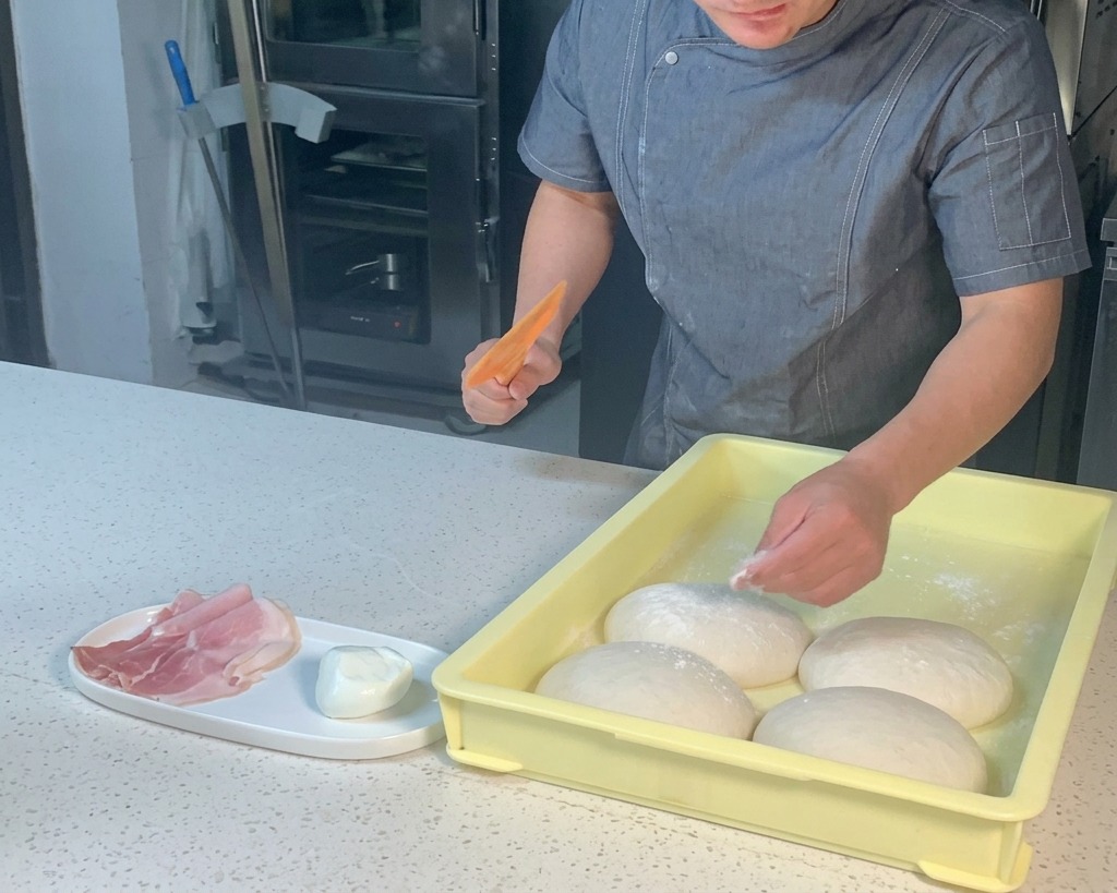 Uno chef usa un tarocco di plastica arancione per sollevare un panetto di pizza da un vassoio giallo.